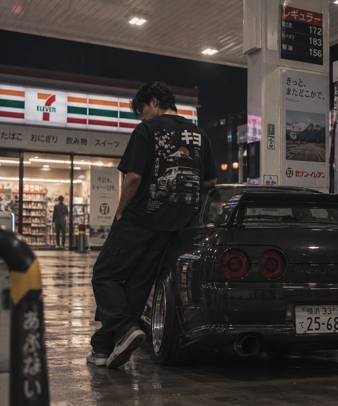 Model wearing a black Kiyo graphic tee beside a Nissan Skyline R32 at a Japanese petrol station